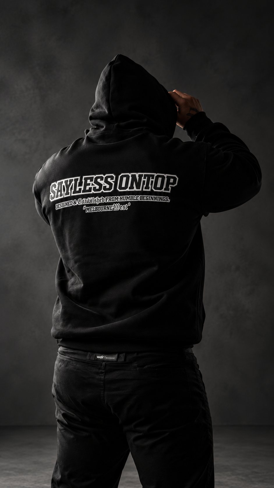 Back of a black SAYLESS ONTOP hoodie, hood up, shot in a dark studio
